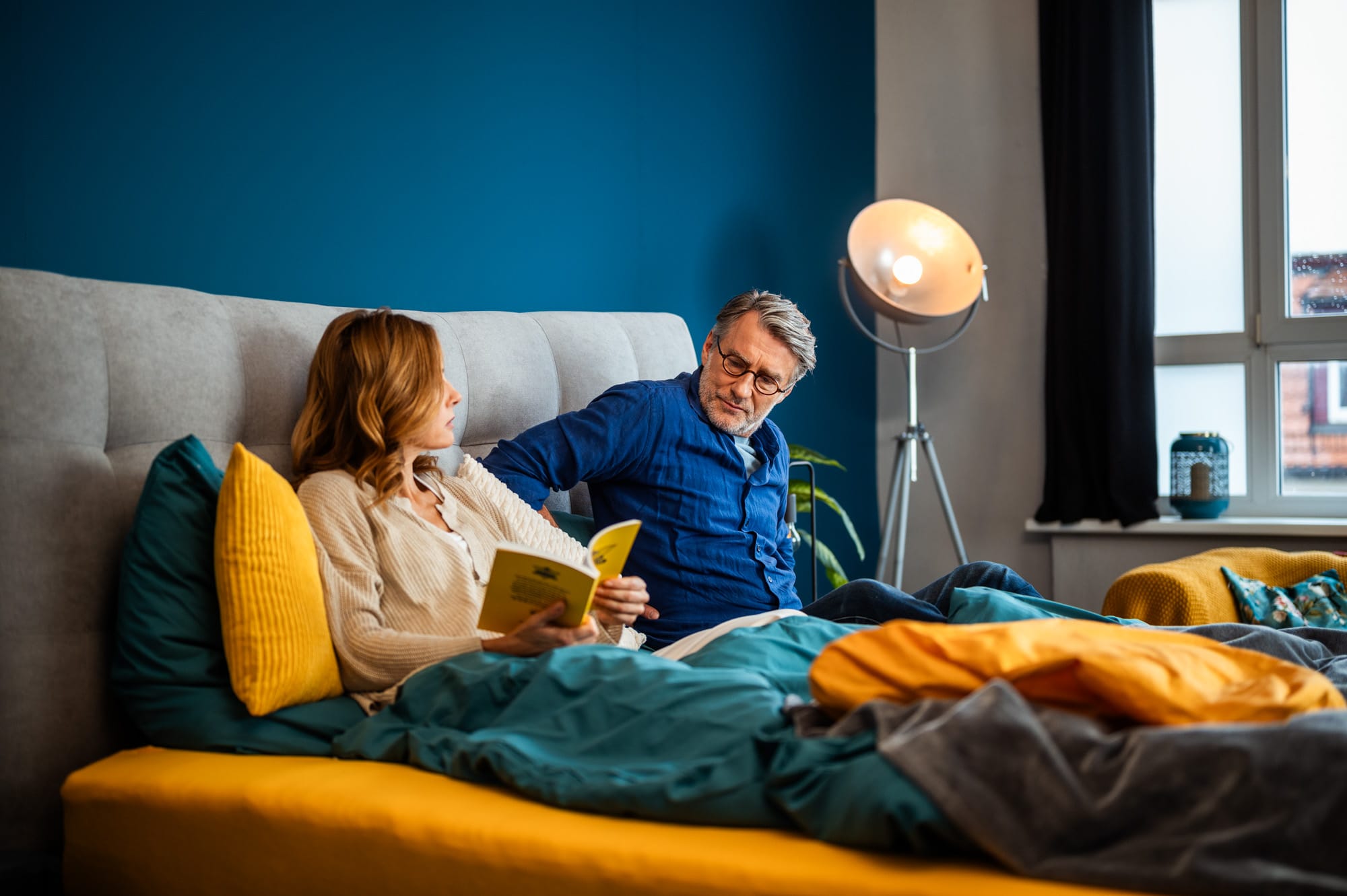 Mann und Frau sitzen gemeinsam im Bett mit Kopfstütze und Kissen und lesen entspannt.