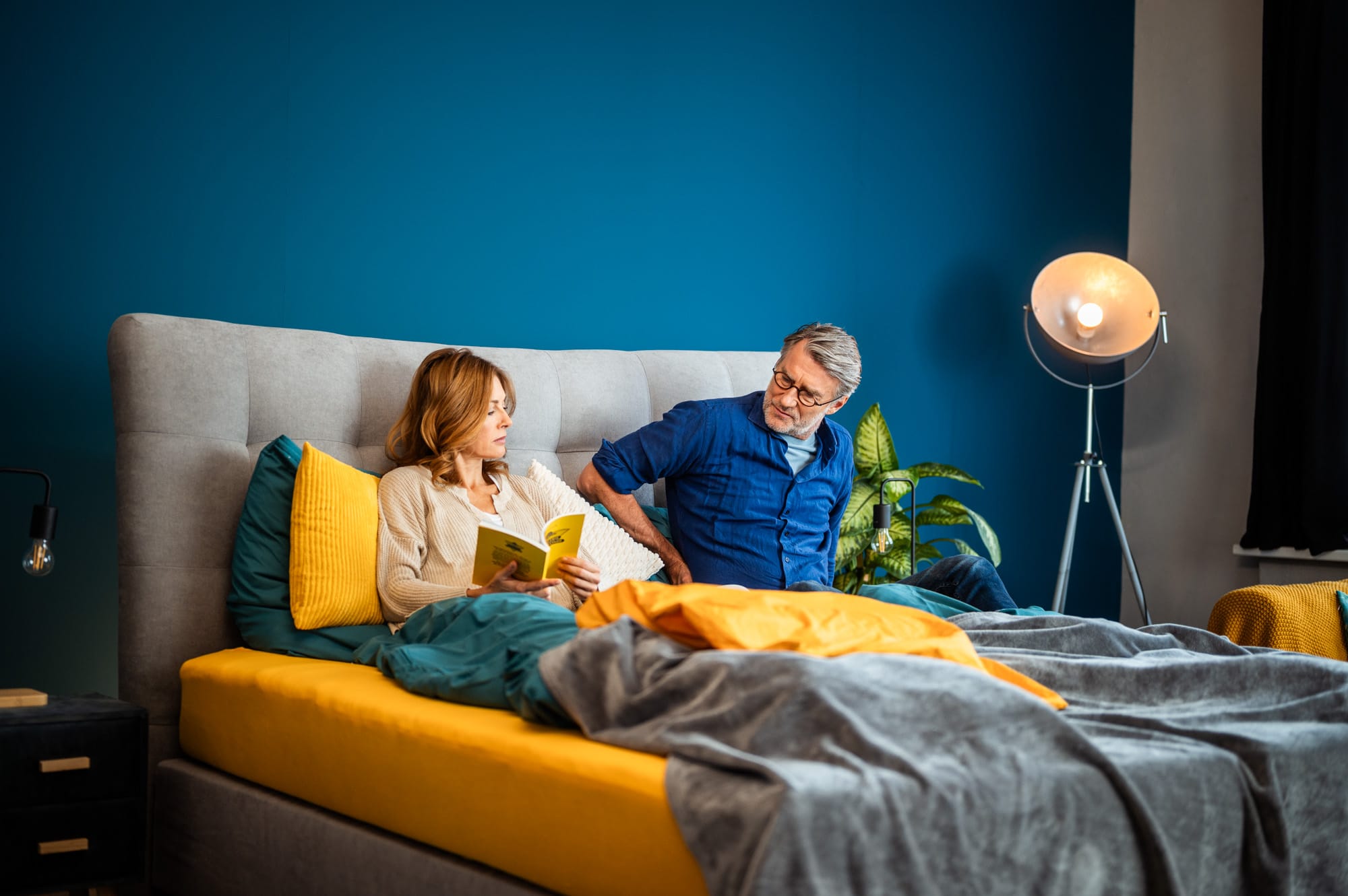 Mann sitzt mit schmerzverzerrtem Gesicht auf einem Bett mit dem CosyLift, während eine Frau daneben liest